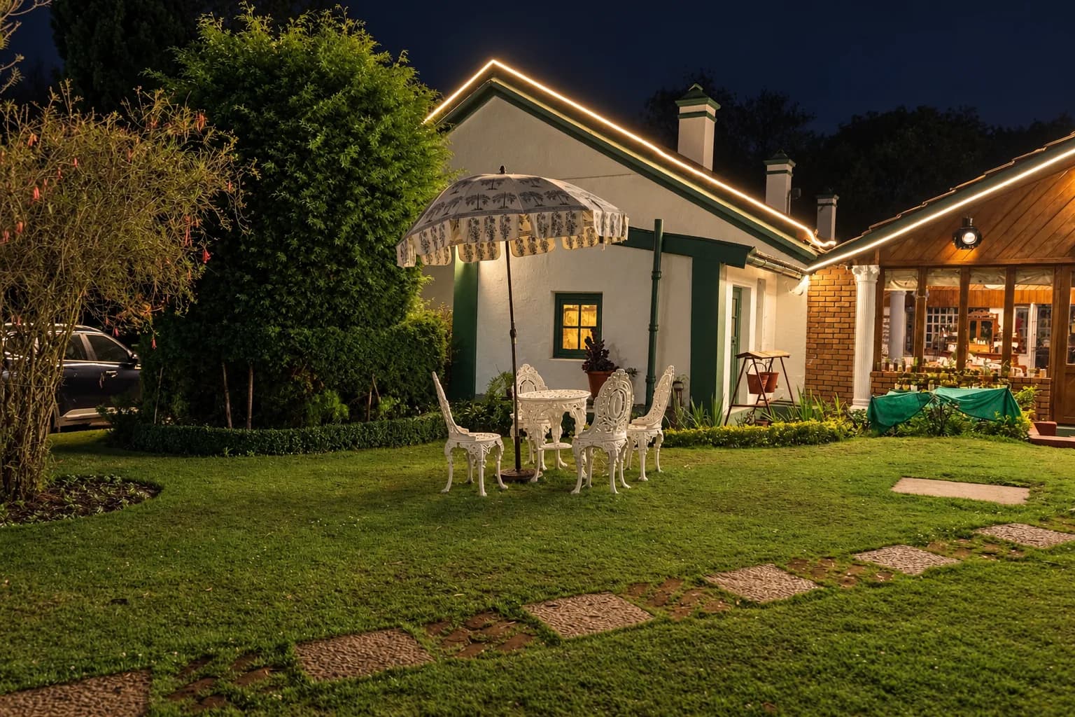 The house at dusk, windows lit, a parasol and white chairs on the lawn
